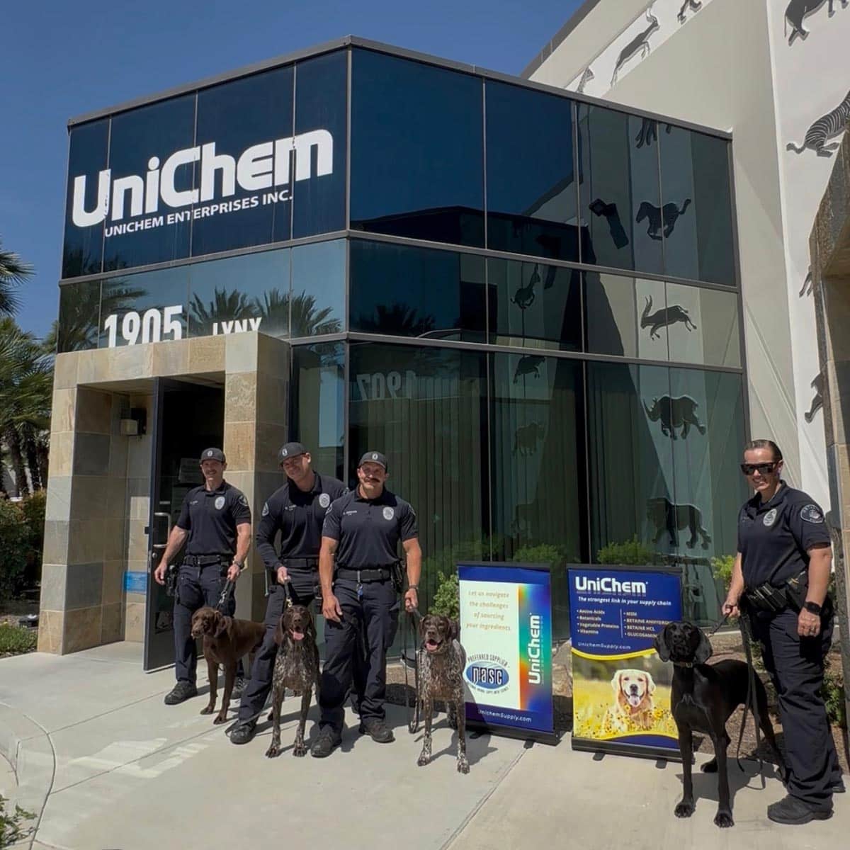 Ontario Police Department of California officers with bomb-sniffing dogs posing in front of a Unichem and NASC posters.